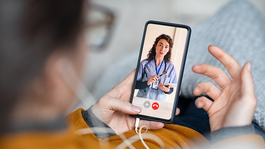 Eine Person, die ein Smartphone in der Hand hält, führt einen Videotelefonanruf an einen Arzt, der einen Kittel und ein Stethoskop trägt und ein Klemmbrett in der Hand hält. Die Person scheint bequem zu sitzen, trägt einen Pullover und Kopfhörer.