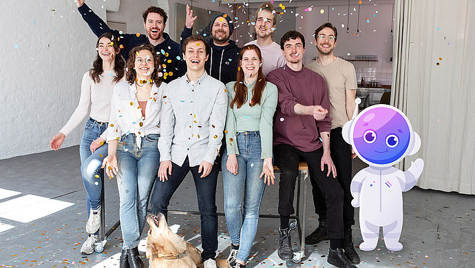 Eine Gruppe von acht lächelnden Menschen und ein Golden Retriever posieren in einem Raum, in dem buntes Konfetti herabfällt. Rechts daneben winkt ein freundlicher lila Cartoon-Roboter. Die Atmosphäre ist hell und leger.