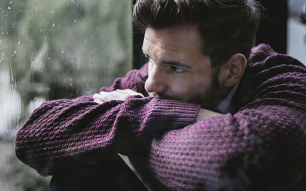 Ein Mann in einem kastanienbraunen Pullover stützt seine Arme auf eine Fensterbank und blickt an einem regnerischen Tag nachdenklich nach draußen. Am Fenster sind Regentropfen zu sehen, und die Szene wirkt düster und nachdenklich.