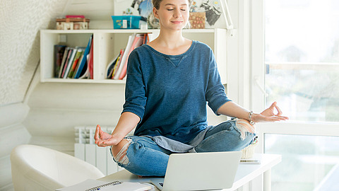 Eine Person sitzt im Schneidersitz auf einem Schreibtisch vor einem Laptop, die Augen sind geschlossen und die Hände ruhen in Meditationshaltung auf den Knien. Im Hintergrund sind Bücher und Regale zu sehen, und das Sonnenlicht erfüllt den Raum.