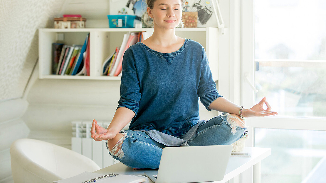 Eine Person sitzt im Schneidersitz auf einem Schreibtisch vor einem Laptop, die Augen sind geschlossen und die Hände ruhen in Meditationshaltung auf den Knien. Im Hintergrund sind Bücher und Regale zu sehen, und das Sonnenlicht erfüllt den Raum.