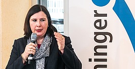 Eine Frau mit schulterlangem, dunklem Haar spricht in ein Mikrofon und gestikuliert dabei mit der Hand. Sie trägt einen dunklen Blazer und einen gemusterten Schal und steht neben einem vertikalen Schild mit dem teilweise sichtbaren Wort „Inninger“.