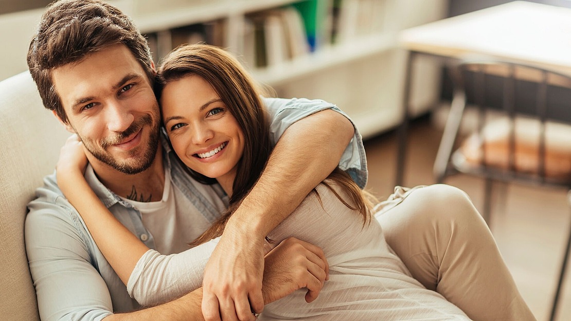 Ein lächelndes Paar sitzt eng beieinander auf einem Sofa, wobei die Frau ihre Arme um den Mann schlingt. Sie wirken glücklich und entspannt in einem gemütlichen, gut beleuchteten Raum mit Regalen im Hintergrund.