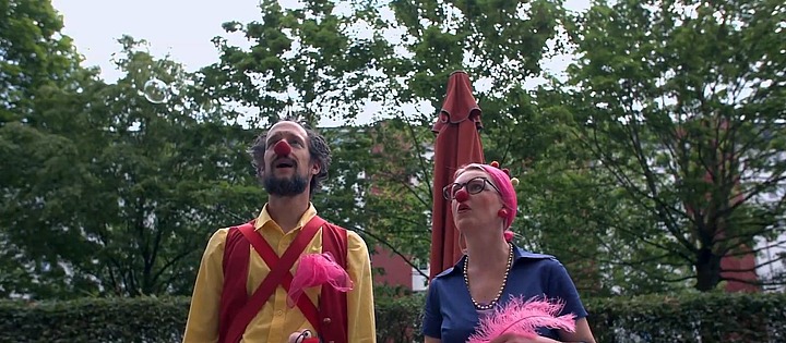 Zwei als Clowns verkleidete Erwachsene, jeder mit einer roten Nase, stehen im Freien vor Bäumen und einem Regenschirm. Einer trägt ein gelbes Hemd mit roten Hosenträgern, der andere ein blaues Kleid und ein rosa Kopftuch. Beide halten rosa Federrequisiten in der Hand.