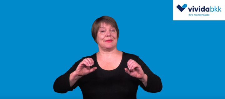 Eine Frau mit kurzen braunen Haaren, die ein schwarzes Oberteil trägt, gestikuliert mit ihrer rechten Hand vor einem blauen Hintergrund. In der rechten oberen Ecke erscheint das Logo von vivida bkk.