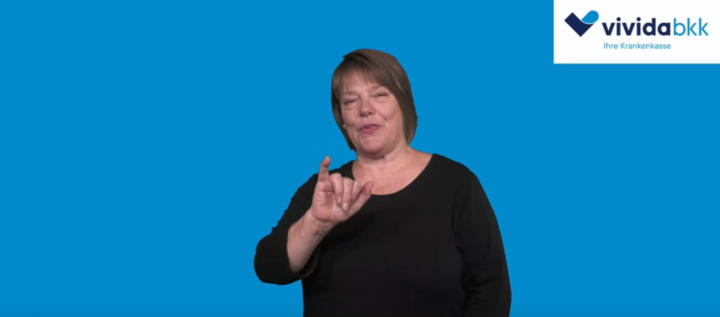 Eine Frau mit kurzen braunen Haaren, die ein schwarzes Oberteil trägt, gestikuliert mit ihrer rechten Hand vor einem blauen Hintergrund. In der rechten oberen Ecke erscheint das Logo von vivida bkk.