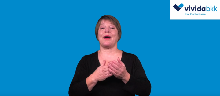 Eine Frau mit kurzen braunen Haaren, die ein schwarzes Oberteil trägt, verwendet Zeichensprache vor einem einfarbig blauen Hintergrund. Das Logo der Krankenkasse vivida bkk erscheint in der oberen rechten Ecke.